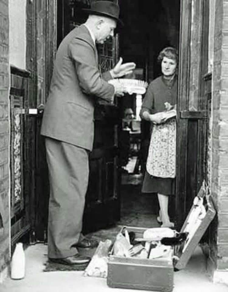 A black and white photo of a door-to-door salesman speaking to a woman in her doorway